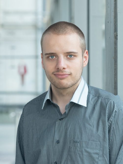 Das Bild zeigt eine Person vor einem neutralen Hintergrund. Die Person schaut authentisch in die Kamera, das Bild ist bewusst ohne KI erstellt.